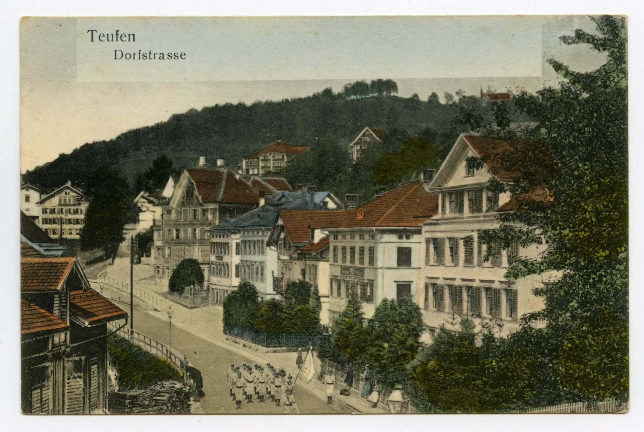 marschierende Vereinsgruppe (Turner?) in weissem Dress, mit Fähnrich und Zaungästen. Zweites Haus v.r.: „Gasthaus Ochsen“. Markanter Brennholz-Stapel links unten (vgl. Hc.20-20)