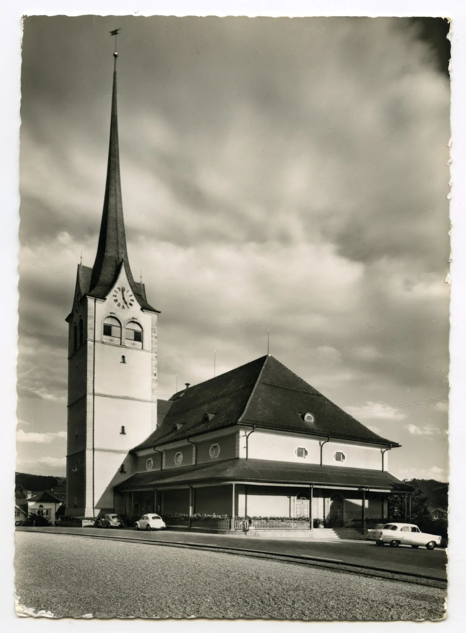 vom Vorplatz des alten Bahnhofs gesehen, mit Bahngeleise. Drei parkierte Autos, davon 2 VW-Käfer entlang der Kirche