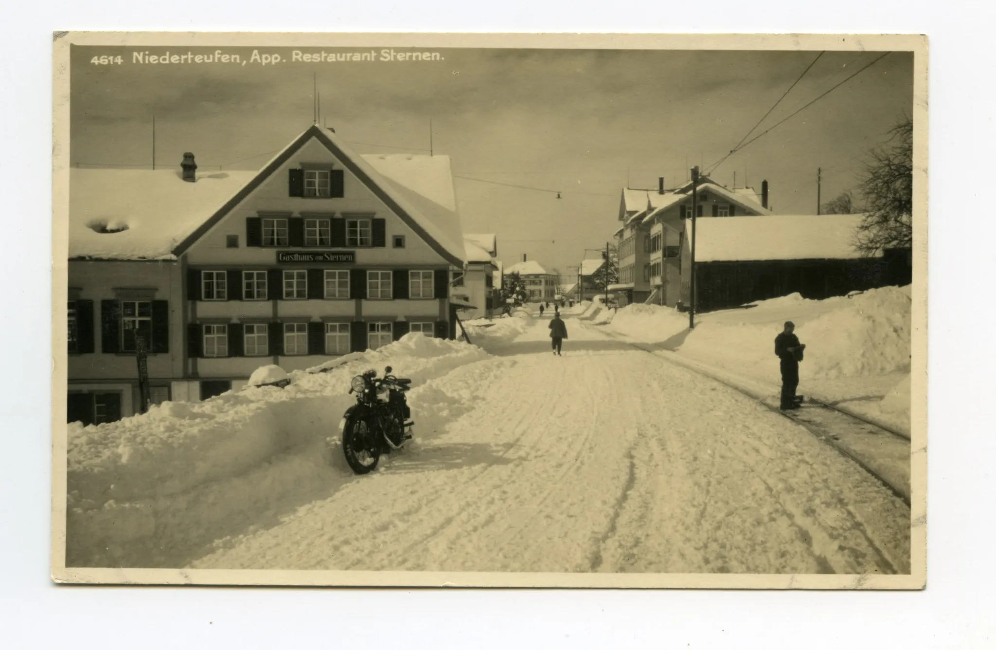 Ansicht aus Osten mit Motorrad, Bahnschiene und Personen auf der schneebedeckten Strasse