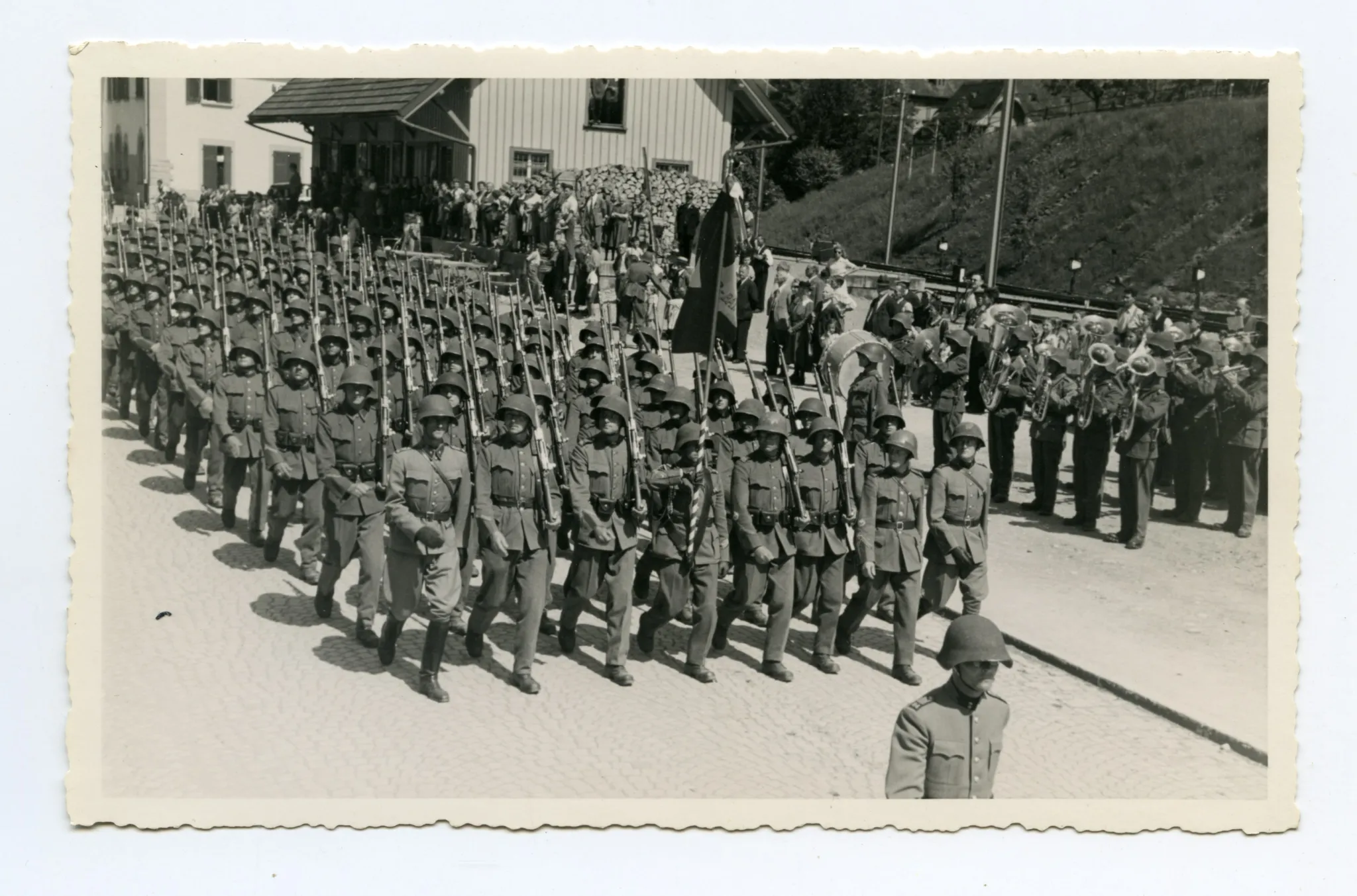 Soldaten (Kompanie) marschieren auf der Strasse (Ebni) Richtung Zeughaus, daneben Militärkapelle vor den Bahnschienen
