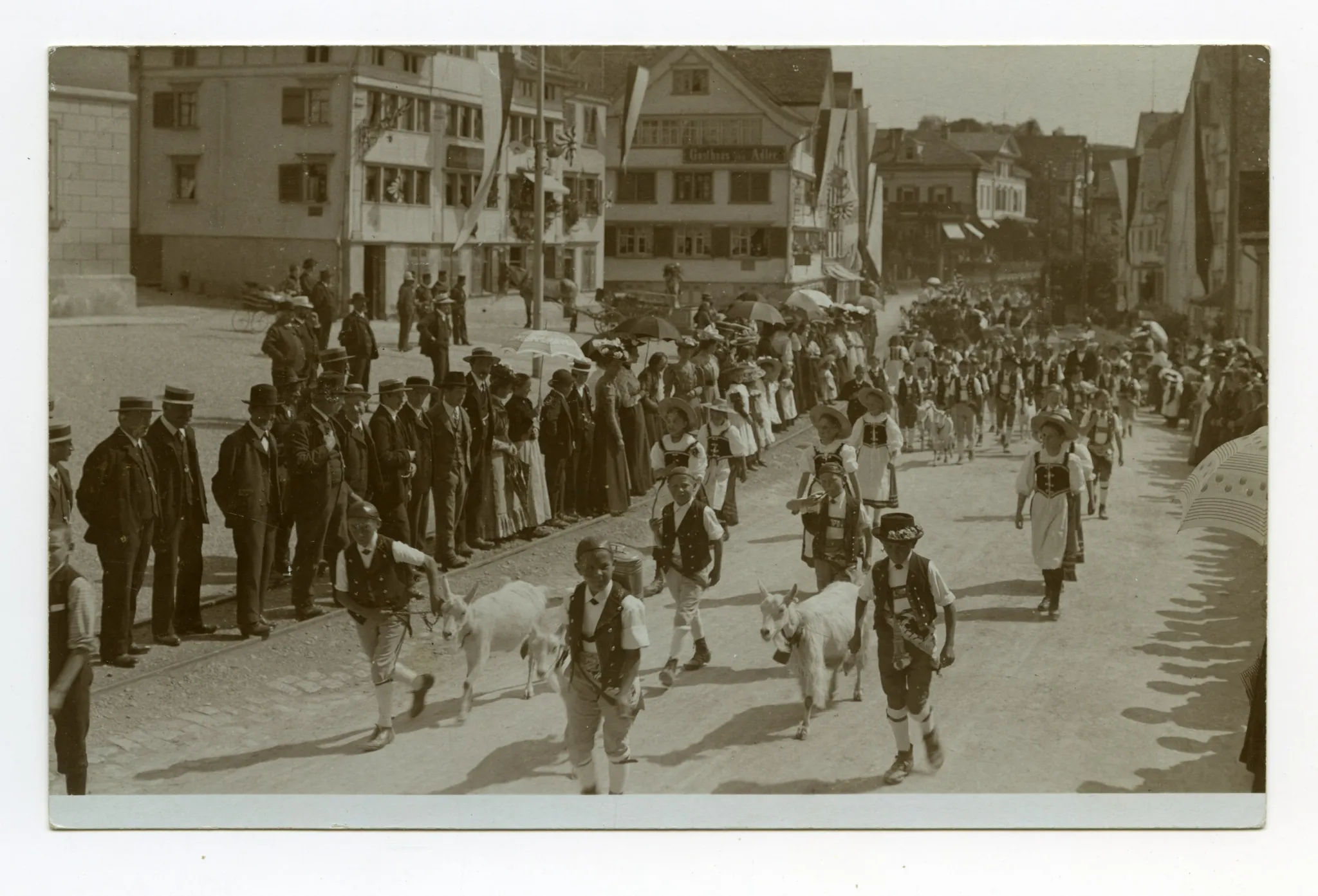 Knaben in Sennentracht und Ziegen, sowie Mädchen in Tracht laufen vor der Häuserzeile mit Gasthaus Anker, Gasthaus zum Adler und Villa Roth, Zuschauer stehen auf der Bahnschiene