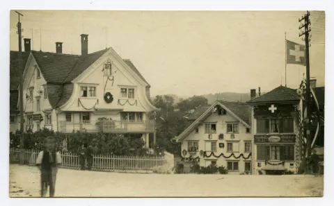 Blick zu Einlenker Hechtstrasse, Hotel Hecht mit Schweizer Fahne und –Wappen an Fassade, Kornhändlerhaus. Buben und Männer auf der Strasse. Häuser mit Blumenranken und Fähnlein verziert