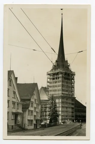 eingerüstet (Holzgerüst bis Höhe Turmuhr), von der Dorfstrasse Ost gesehen. Bauarbeiter mit weissen Kurzarm-Hemden, am Fuss des Turms sowie auf dem Gerüst