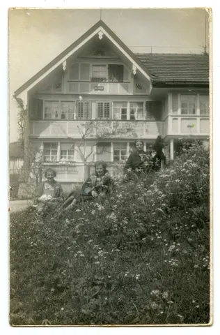 Ansicht mit Hausbewohnerinnen (?) sitzend auf der Wiese vor dem Haus