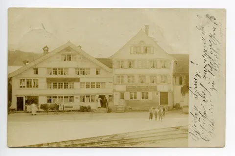 Metzgerei im Februar 1980 eingegangen, Drogerie 1966 eingegangen, Frontansicht mit Personen davor, Ochse wird vor der Metzgerei gehalten, Eisenbahnschienen im Vordergrund sichtbar, Drogerie Schlatter mit im Bild