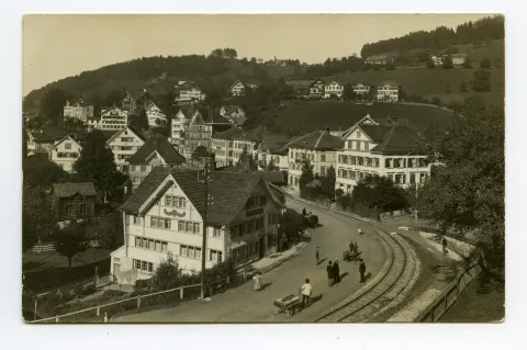 Ansicht aus Südosten mit Hauptstrasse-Verlauf, Bahngleis, Häuserzeile mit Drogerie Alpina, Restaurant Metzgerei Ochsen, Personen auf der Strasse, Blick Richtung Frohe Aussicht