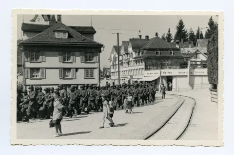 Soldaten marschieren auf der Hauptstrasse Richtung ZeughausBahnhofskreuzung, im Hintergrund Bekleidungsgeschäft F. Wetter