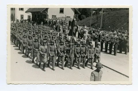 Soldaten (Kompanie) marschieren auf der Strasse (Ebni) Richtung Zeughaus, daneben Militärkapelle vor den Bahnschienen