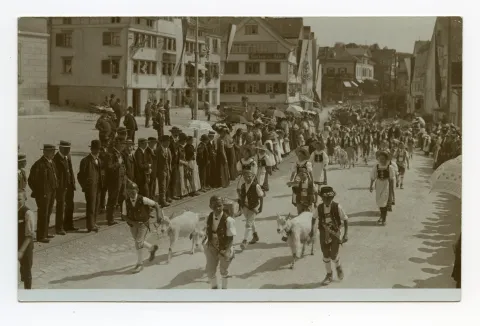 Knaben in Sennentracht und Ziegen, sowie Mädchen in Tracht laufen vor der Häuserzeile mit Gasthaus Anker, Gasthaus zum Adler und Villa Roth, Zuschauer stehen auf der Bahnschiene