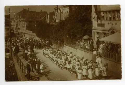 Gruppe von Mädchen mit Musikanten im Anschluss läuft am Anfang der Speicherstrasse, mit Schaulustigen am Strassenrand und vor dem Restaurant Blume