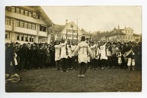 „Vorführung“ gesäumt von Schaulustigen, ein Kind verkleidet als Harlekin, im Hintergrund Café Spörri
