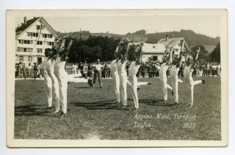 neun Turner synchron beim Turnen einer Übung auf dem Zeughausplatz, im Hintergrund Kinderkurhaus Sanitas
