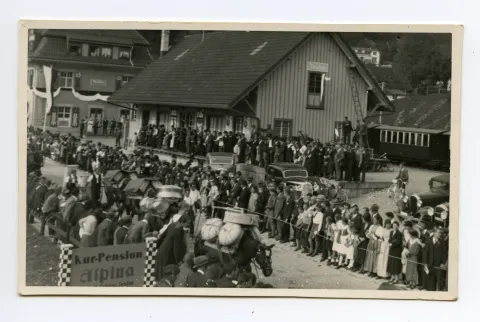 Saumpferde beladen mit Sennengeschirr gefolgt von Sennen mit Tanse auf dem Rücken vor dem Güterschuppen