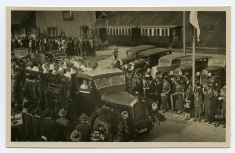 Lastwagen (Berna) mit Jugendharmonikerorchester, Sänger und Sängerinnen auf dem offenen Wagen, im Hintergrund Güterschuppen