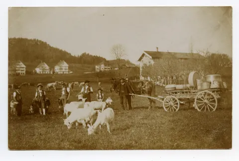 in Tracht und Fuhrwerk, beladen mit Sennengeschirr auf der Wiese im Landhaus, im Hintergrund Häuserfront an der Speicherstrasse